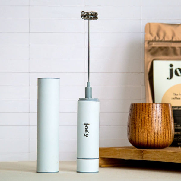 white froth'y hand frother next to a wooden mug and a bag of joe'y coffee alternative